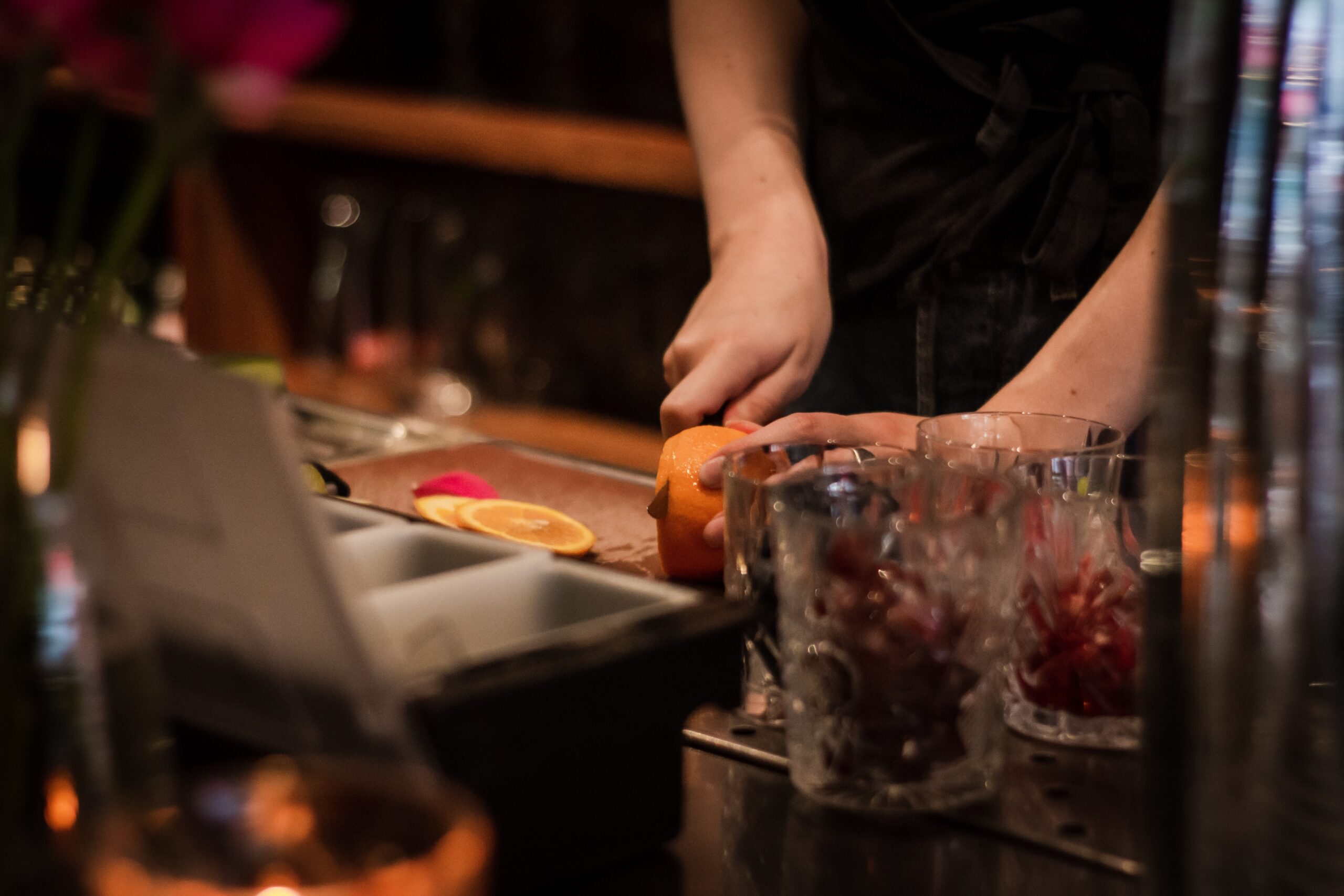 A selective focus shot of bartender slicing oranges on a tray near glasses